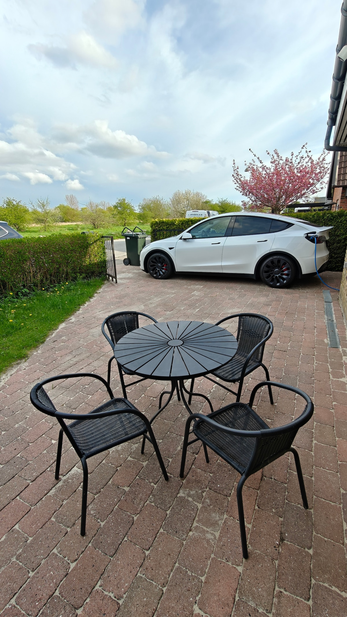 An outdoor seating area features a round table surrounded by four black chairs on a brick patio. A white car is parked nearby, with greenery and flowering trees providing a natural backdrop. The sky is partly cloudy, adding to the inviting atmosphere.