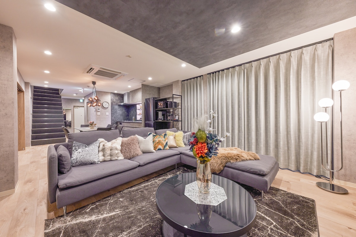A spacious living area features a large, contemporary sectional sofa adorned with various patterned cushions. A glass coffee table sits atop a stylish area rug. Soft lighting is provided by a floor lamp and overhead fixtures, while an expansive curtain gently frames the windows.