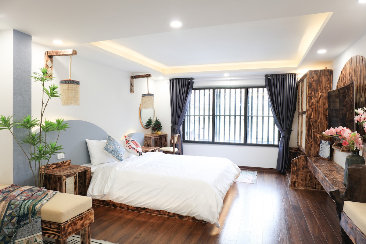 A spacious room displays a large bed with white bedding against a light-colored accent wall. Natural wood furniture, including bedside tables and a bench, complements the warm wooden floor. A window with multiple panes allows natural light, while dark curtains frame the view.
