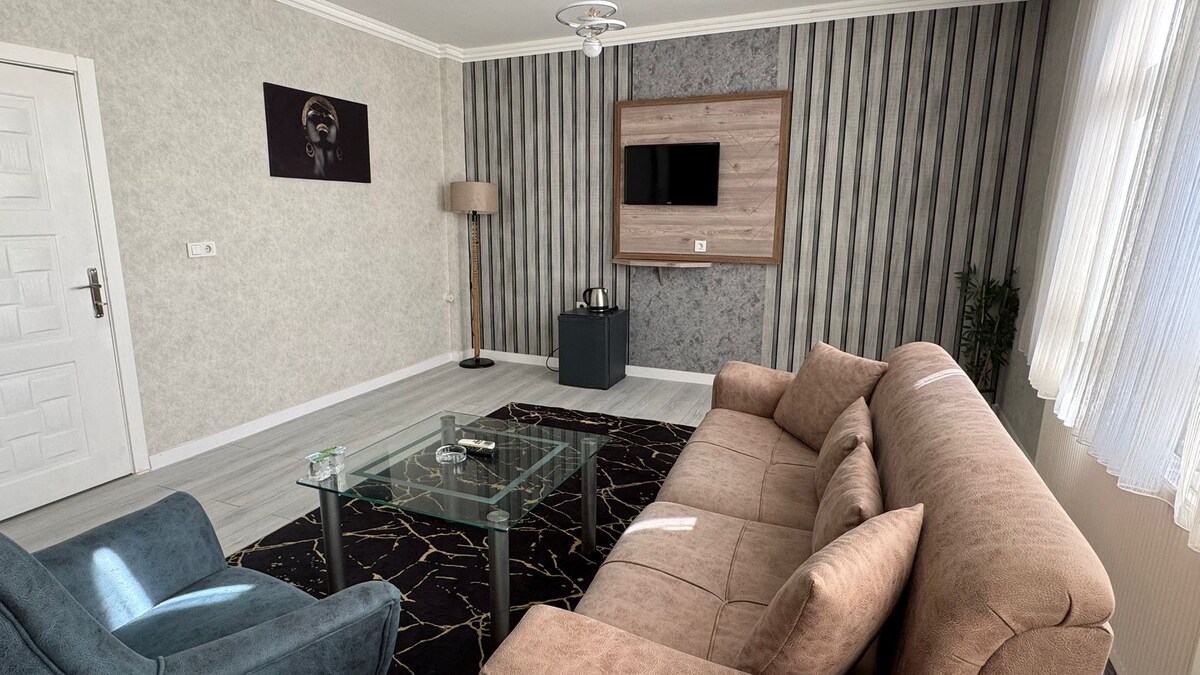 A modern living area features a brown sofa and a blue accent chair, arranged around a glass coffee table. A wall-mounted television is positioned above a wooden panel. Natural light filters through white curtains, enhancing the clean, minimalist design.