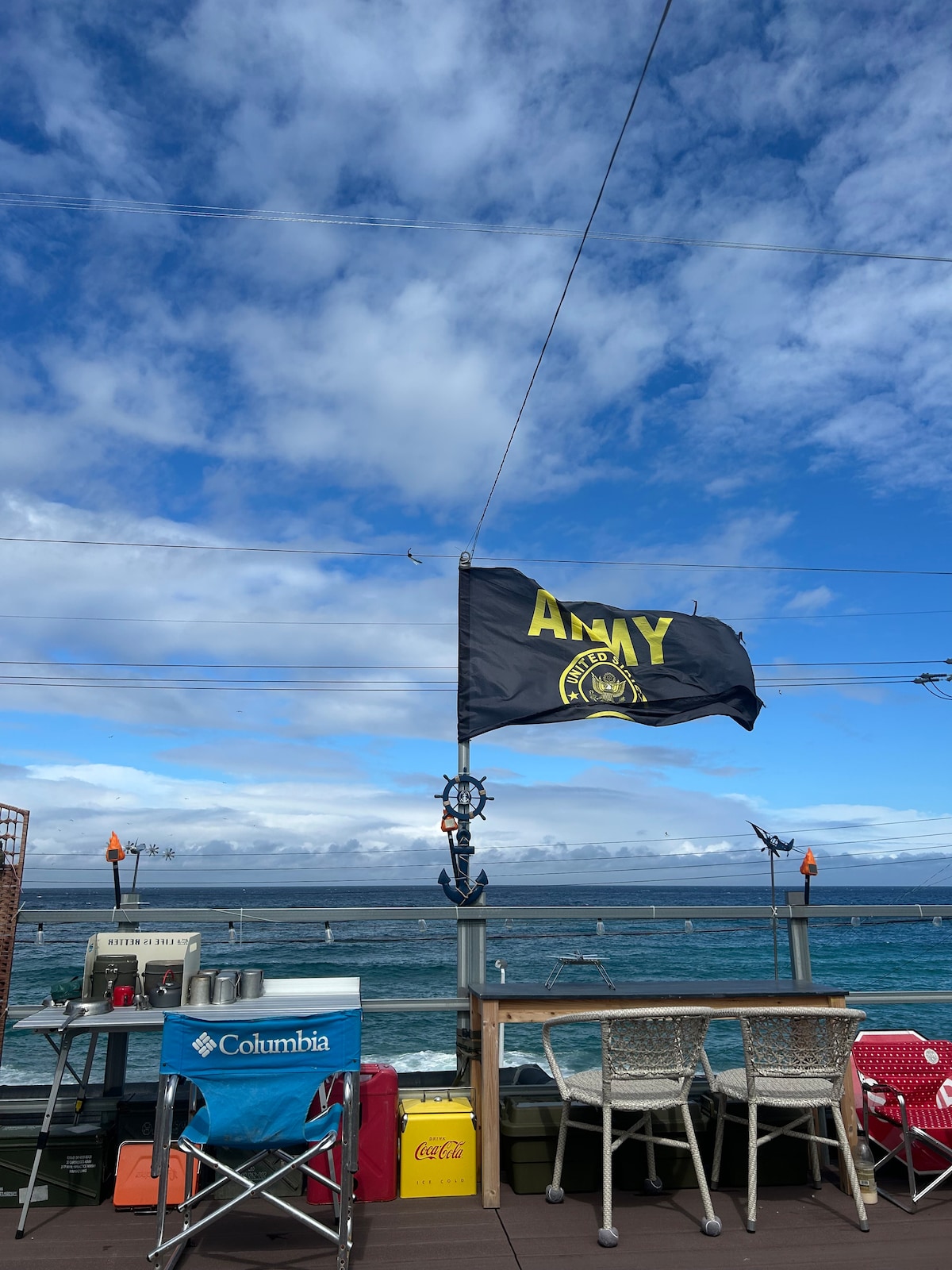 A flag displaying 'AMY' waves gently in the breeze above a patio area. Colorful chairs and tables occupy the space, with a view of the ocean and blue sky filled with white clouds in the background. The natural scenery highlights a relaxed outdoor setting.