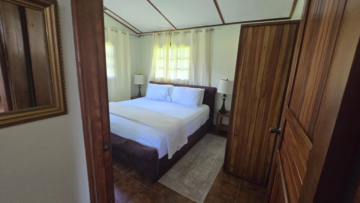 A spacious sleeping area is present, featuring a large bed dressed in crisp white linens. Soft light filters through the sheer curtains, illuminating the room. Wooden doors and a framed mirror add warmth to the natural decor.