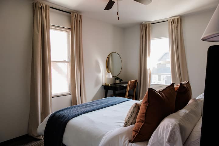 Kitanda kilichotengenezwa vizuri kilicho na mashuka meupe, blanketi la jeshi la wanamaji na mito yenye rangi ya hudhurungi yenye joto. Mapazia ya beige yanafunika madirisha mawili, yakiruhusu mwanga wa asili kuingia. Ubatili mdogo mweusi ulio na kioo cha mviringo na kiti cha fito kimewekwa kwenye kona.