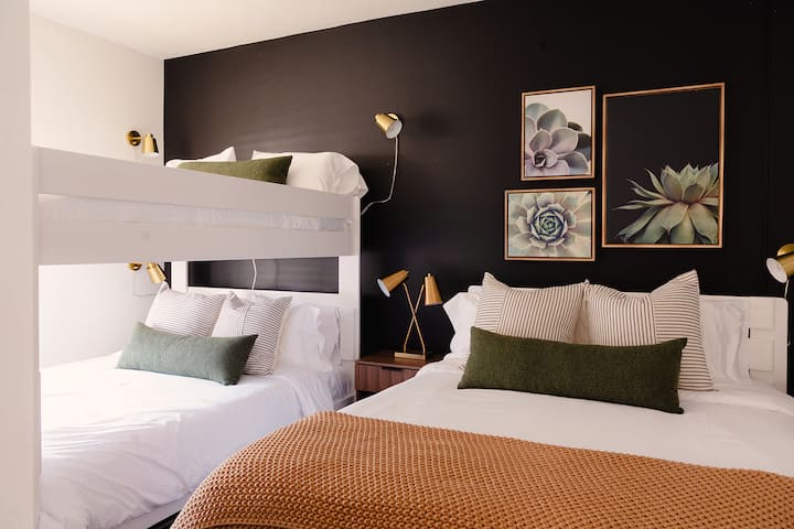 Chumba hiki cha kulala cha kisasa kina ukuta mweusi wenye chapa za kupendeza zenye fremu, ghorofa nyeupe na vitanda vya kifalme vyenye mito yenye mistari na kijani kibichi, taa za dhahabu na blanketi la rangi ya hudhurungi.