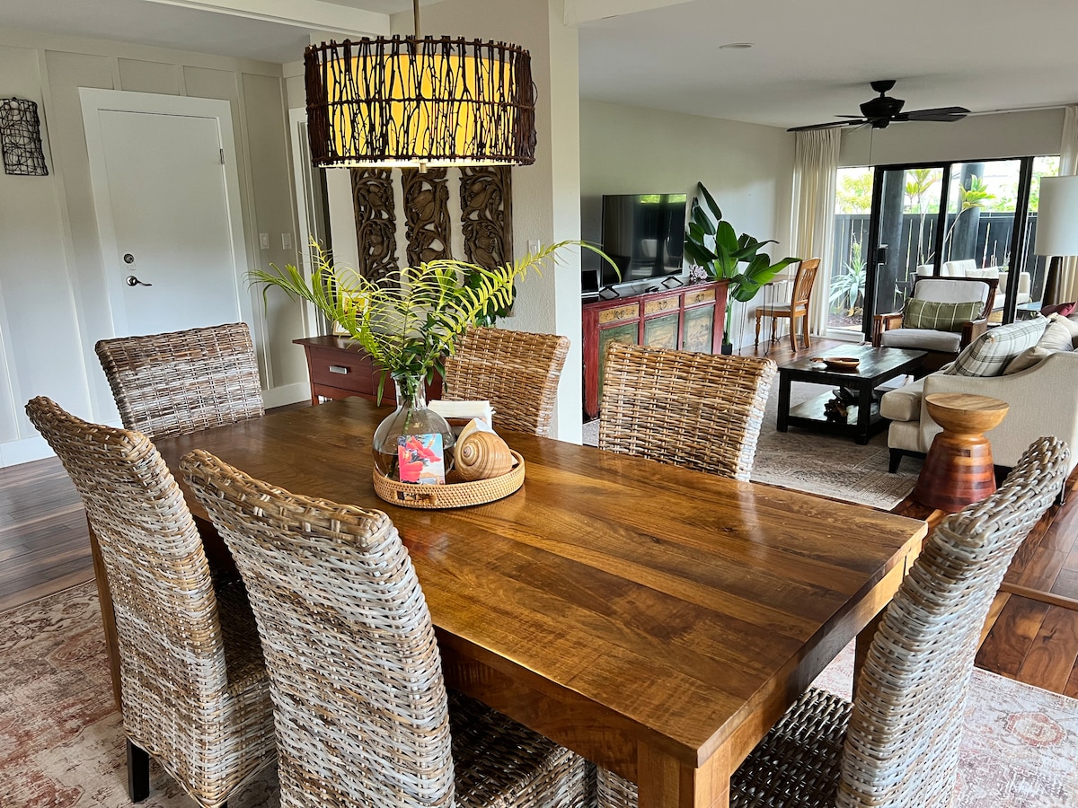 A large wooden dining table is surrounded by six woven chairs, setting a welcoming space for meals. Above, a textured pendant light provides ambient illumination. In the background, a living area is visible, featuring a television and tropical plants, enhancing the inviting atmosphere.