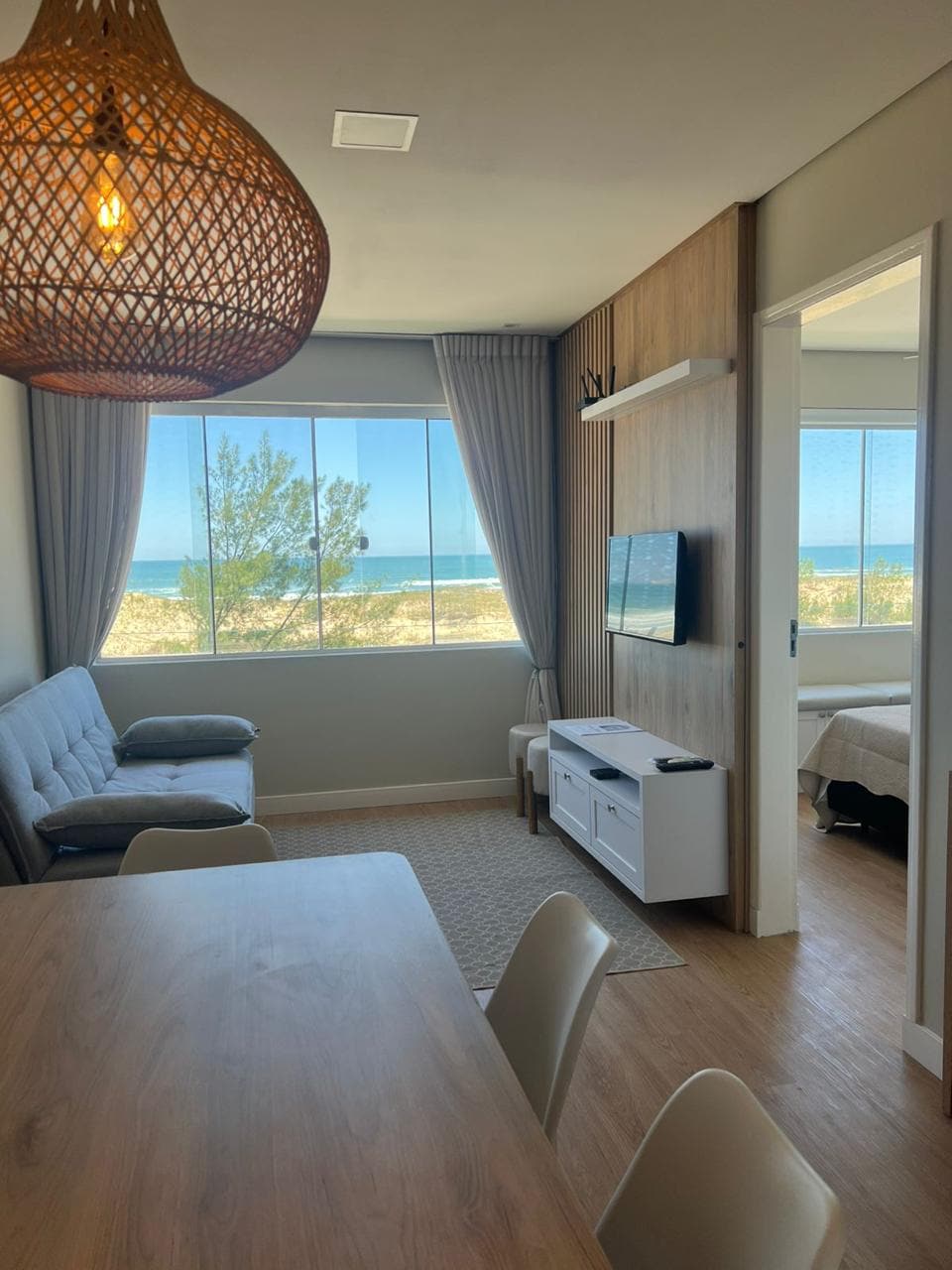 A light-filled living area showcases a comfortable seating arrangement and a wall-mounted television. Large windows provide a view of the ocean, while the wood-paneled accent wall adds warmth. A dining table is present, accompanied by chairs and a woven pendant light above.