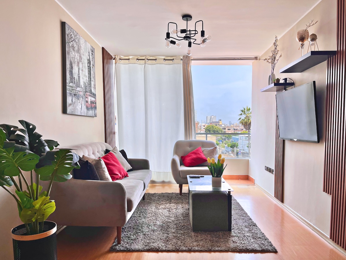 A modern living area features a gray sofa and a comfortable chair, complemented by decorative cushions and a coffee table. Natural light enters through large windows, revealing city views. A vibrant indoor plant adds a touch of greenery, while art adorns the walls.