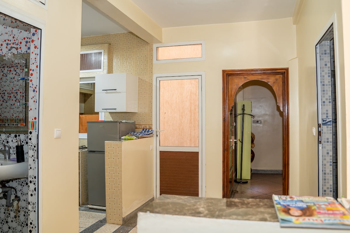 The image captures the view of an open-concept living space, featuring a kitchenette with modern appliances. A door leads to another room, and a cozy seating area is visible on the right. Natural light enters through several windows, creating a bright and inviting atmosphere.