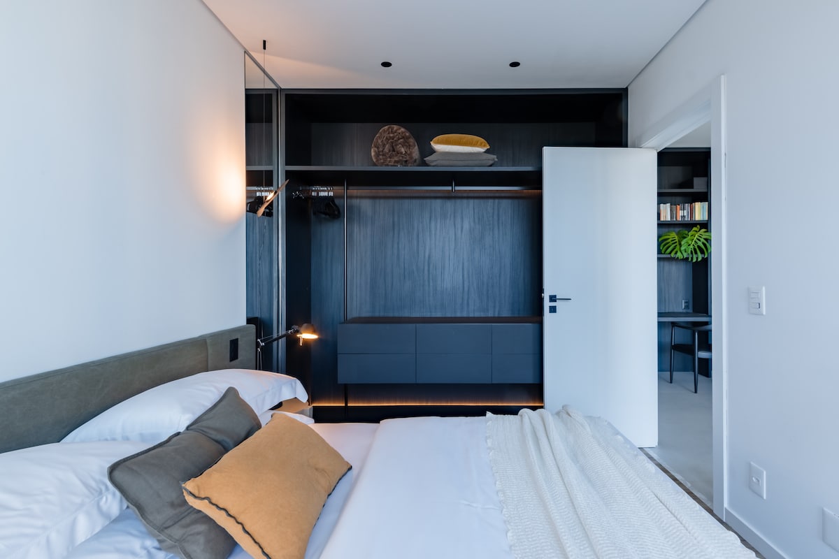 The bedroom features a spacious bed with light bedding and decorative pillows. A sleek closet occupies one wall, providing ample storage. Natural light illuminates the room, highlighting the modern design elements and a decorative item placed above the closet.