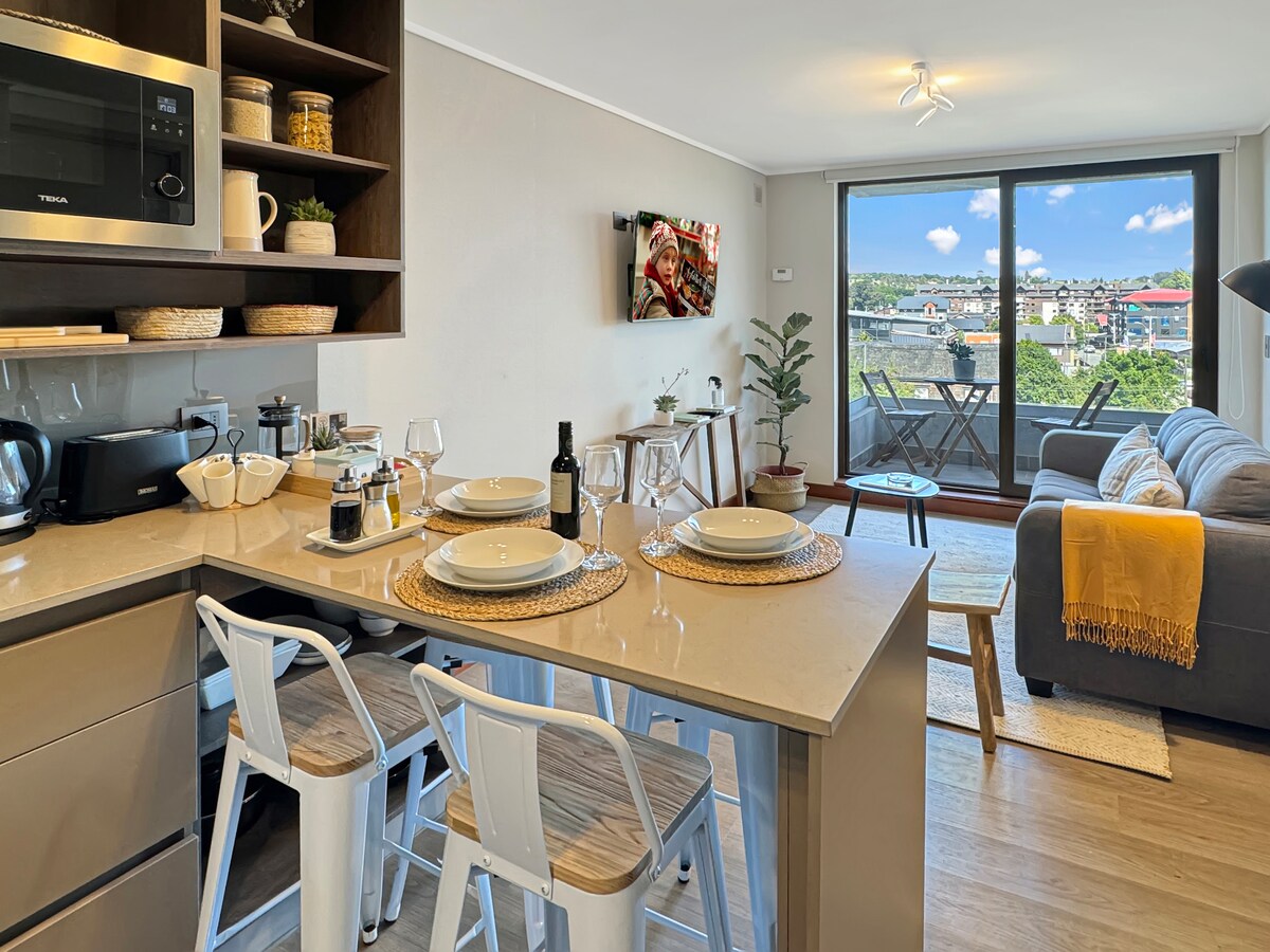 A modern kitchen area is showcased, featuring a well-equipped countertop with dining essentials. Nearby, a cozy living space is highlighted, complete with a comfortable couch and a small table. A large window provides natural light and a view of the outdoor balcony.