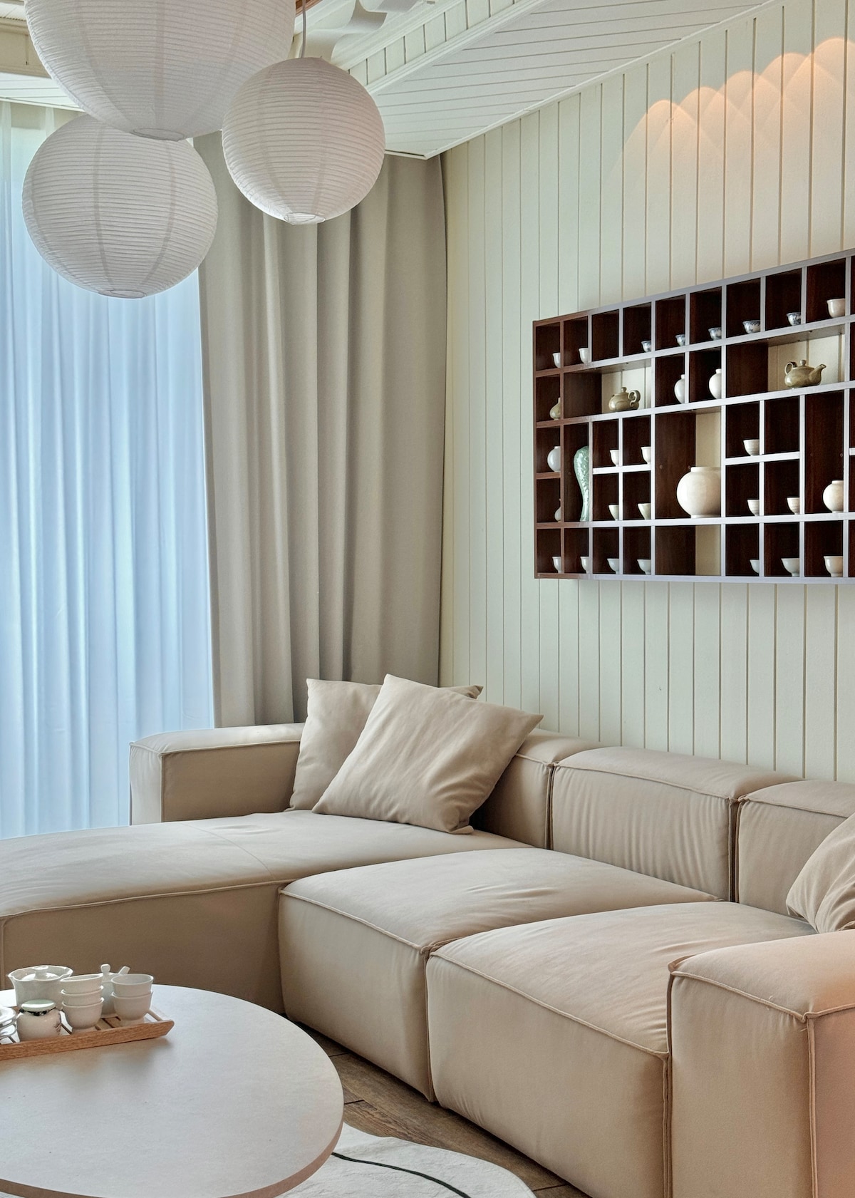 A light and airy living area is presented with a plush sectional sofa adorned with soft neutral cushions. Natural light filters through sheer curtains, creating a warm atmosphere. A decorative shelf holds various ceramics, adding an elegant touch to the space.