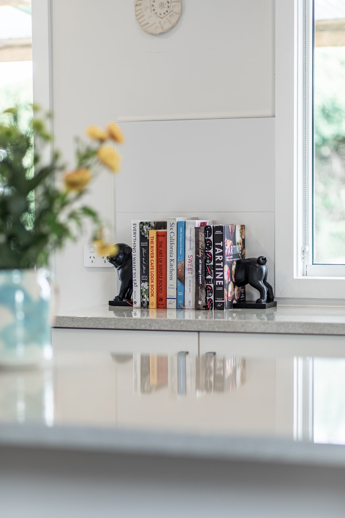 A set of books stands on a shelf, flanked by decorative black dog figurines. Natural light filters through a nearby window, illuminating a small vase of yellow flowers placed beside the books. The countertop showcases a clean and modern aesthetic.