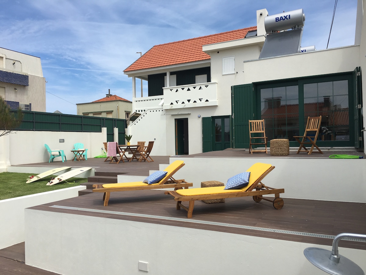 A sun-drenched outdoor area features wooden sun loungers with yellow cushions, positioned on a raised deck. The space is complemented by a dining table and chairs, along with surfboards resting against the wall. A spacious white house with green shutters serves as the backdrop.