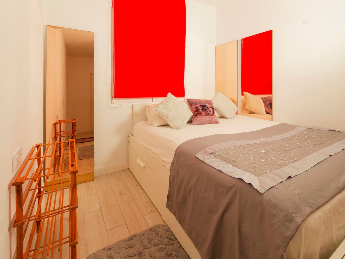 A private room features a spacious bed adorned with decorative pillows and a textured blanket. Two mirrors reflect the light, creating an open feel. Light-colored wooden flooring complements the neutral walls, and a small rack is positioned near the entrance.