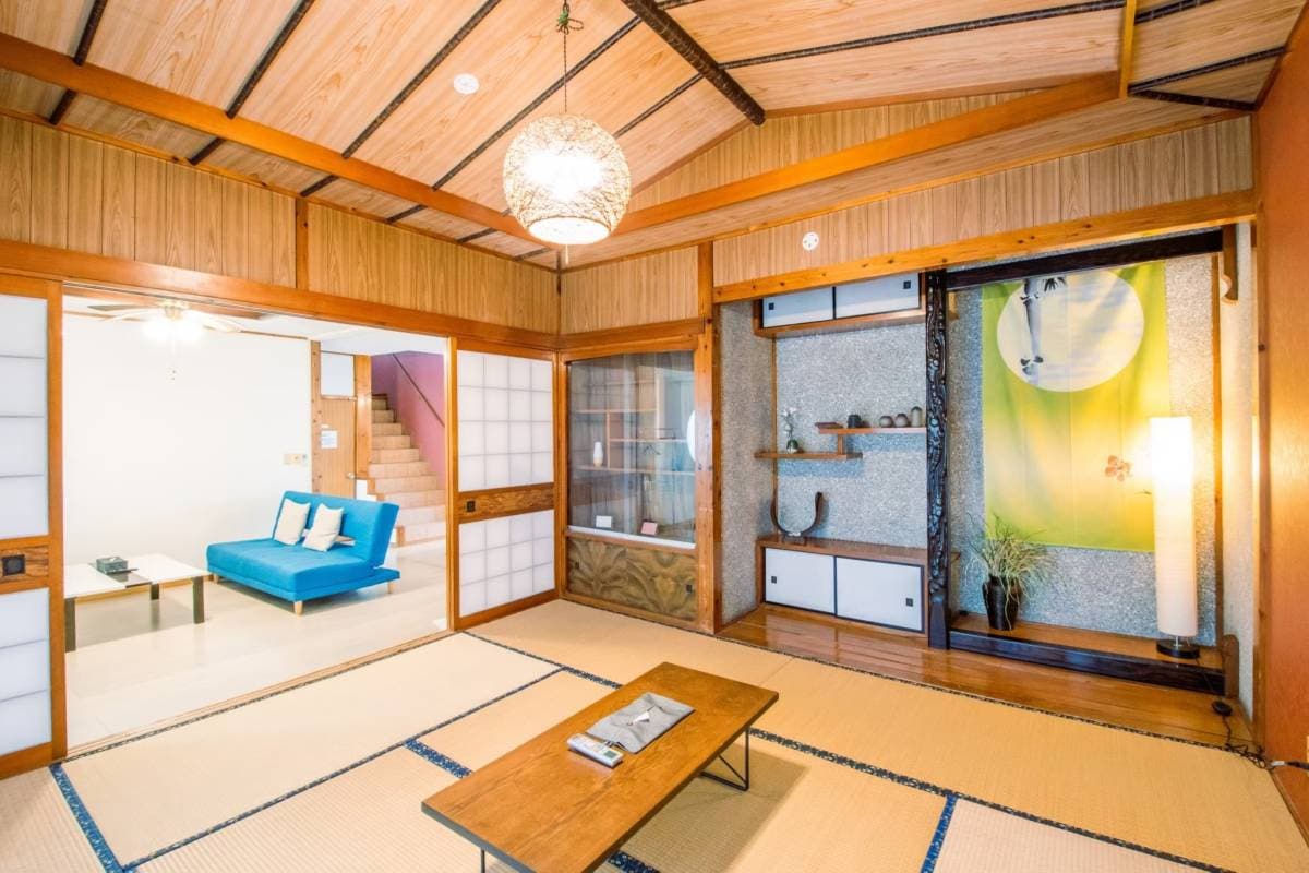 A traditional-style living area features tatami flooring and wooden beams overhead. A low wooden table sits centered on the floor, surrounded by cushions. Sliding doors allow entry to a brightly colored room with a blue sofa. Natural light filters through the windows, enhancing the space.