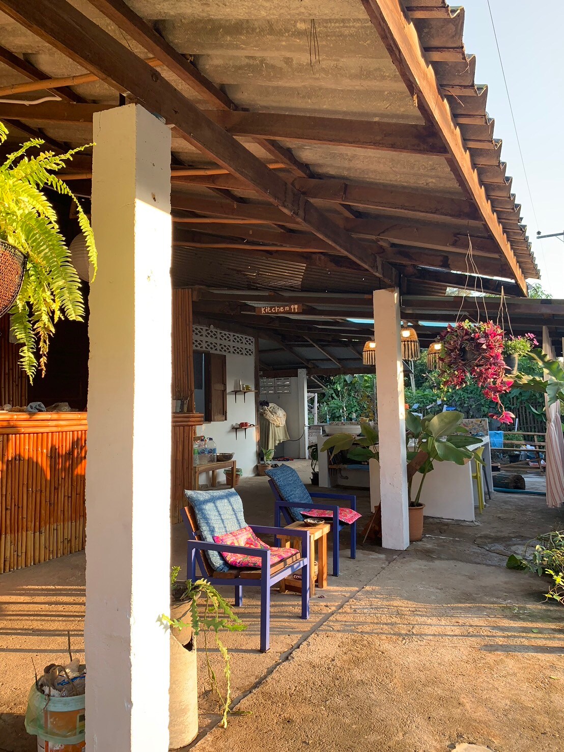 A covered outdoor area features tall wooden pillars and a sloped roof, with seating arrangements comprising colorful chairs. Potted plants and hanging greenery add a touch of nature, while warm sunlight filters through, creating a welcoming ambiance.