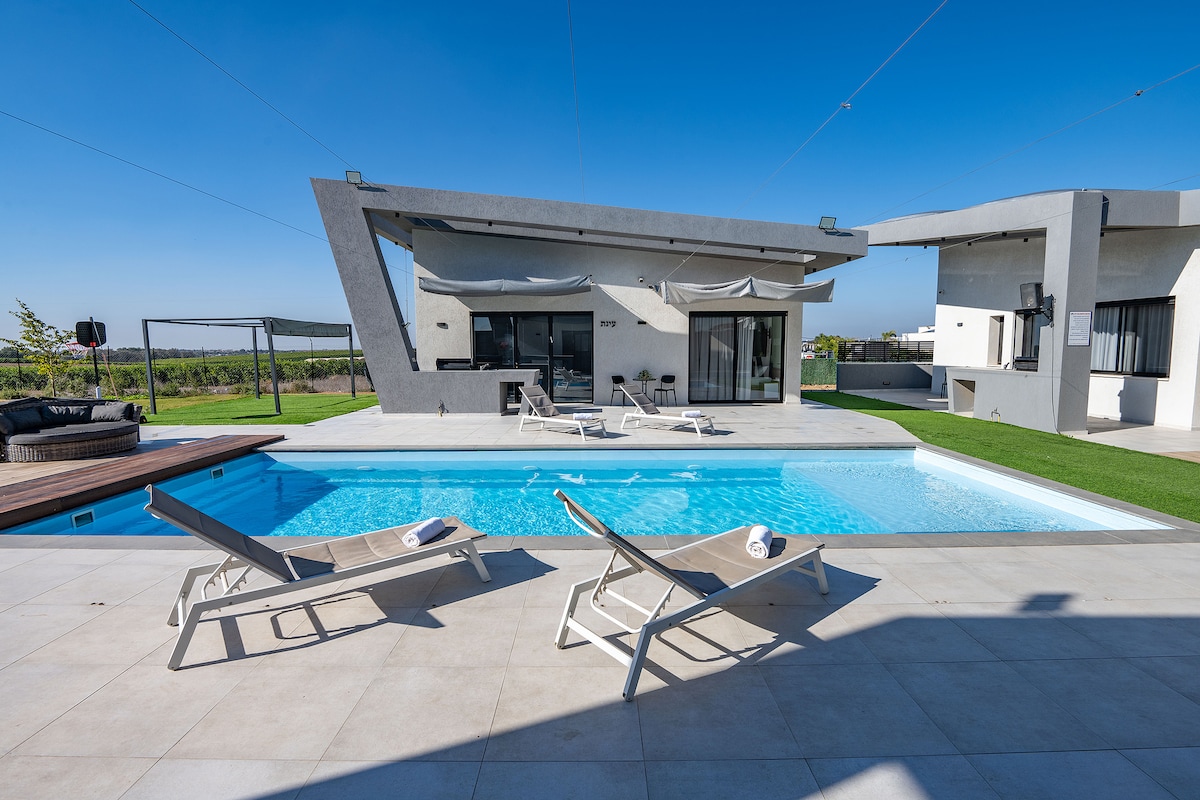 An outdoor area features a rectangular swimming pool surrounded by sun loungers. The modern structure showcases large windows and a shaded deck area. Lush green grass complements the poolside, and the open sky provides a bright and inviting atmosphere.