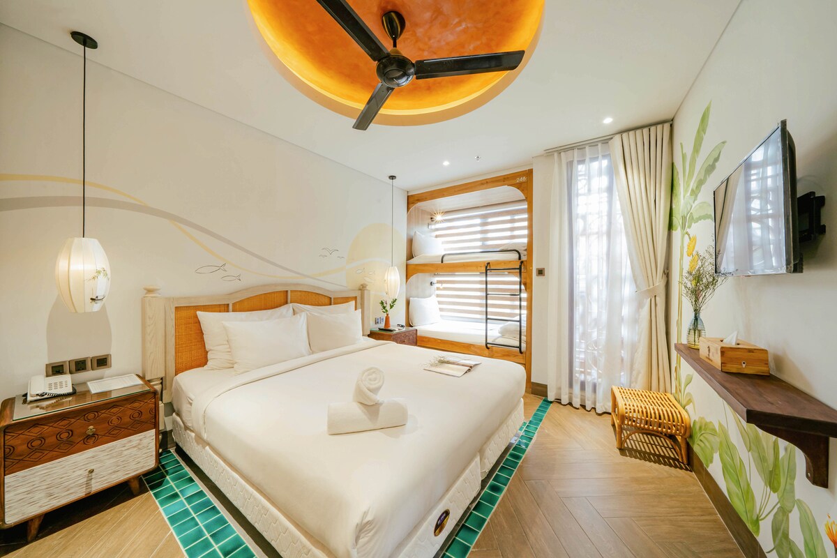 A well-lit bedroom features a spacious bed with crisp white linens and a towel neatly arranged on top. Accent walls are adorned with botanical designs. A flat-screen TV is mounted on the wall, while a stylish fan hangs from the ceiling.