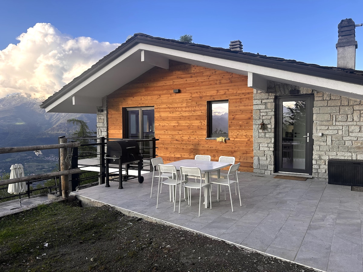 An exterior view of a chalet features a wooden and stone facade, surrounded by the scenic mountains. A spacious patio is equipped with a dining table and chairs, alongside a grill. The space enjoys natural light and protected seating areas, ideal for varying weather.