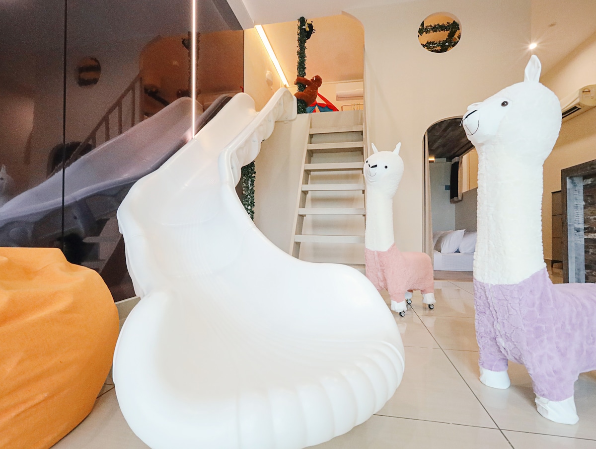 A whimsical indoor area is highlighted by a large, white shell-shaped slide and two plush llama figures, one pink and one white. A staircase leads to the upper level, adding a sense of adventure to the space. Colorful bean bags provide additional relaxation spots.