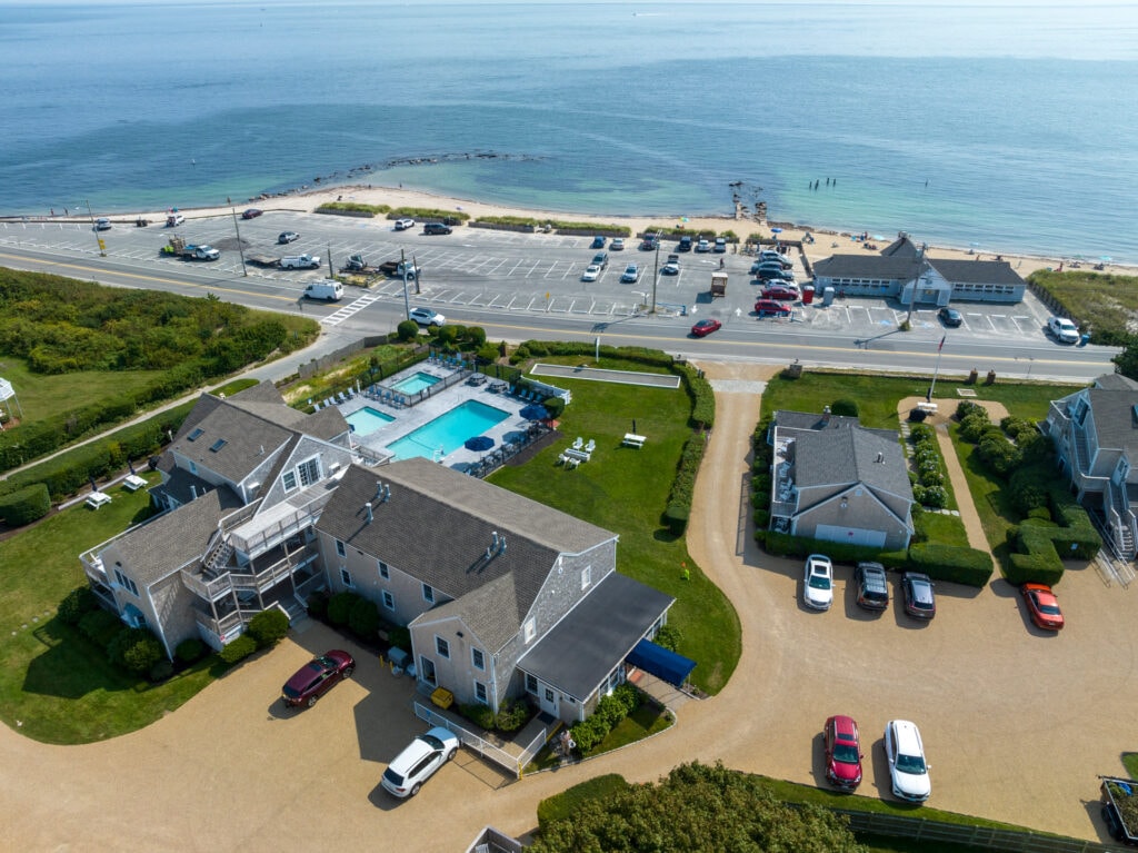 An aerial view captures the beachfront resort with a heated swimming pool centrally located. The neighboring beach and parking area are visible, along with well-maintained greenery surrounding the resort buildings. The calm ocean waters reflect the sky, emphasizing the resort's proximity to the beach.