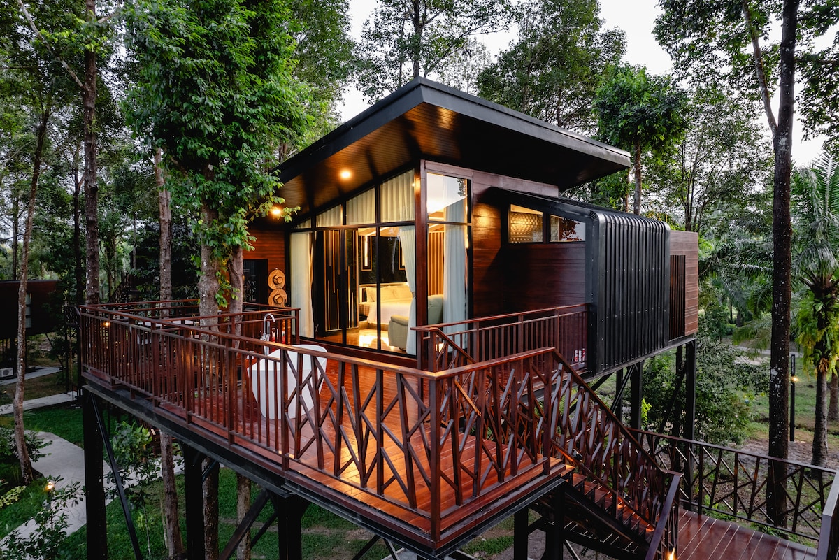 A modern treehouse is nestled among lush trees, featuring a spacious balcony with wooden railings. Large glass windows allow natural light to fill the interior, highlighting the cozy living area and inviting ambiance of the unique accommodation.