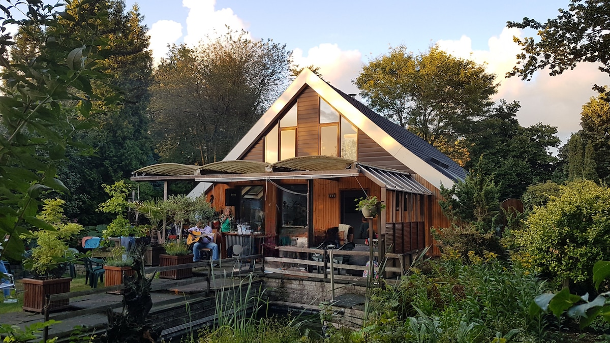 A charming house with a sloped roof is surrounded by lush greenery and outdoor seating areas. Large windows allow natural light to fill the interior. A serene pond is visible in the foreground, complemented by plants and decorative elements, creating a harmonious outdoor space.