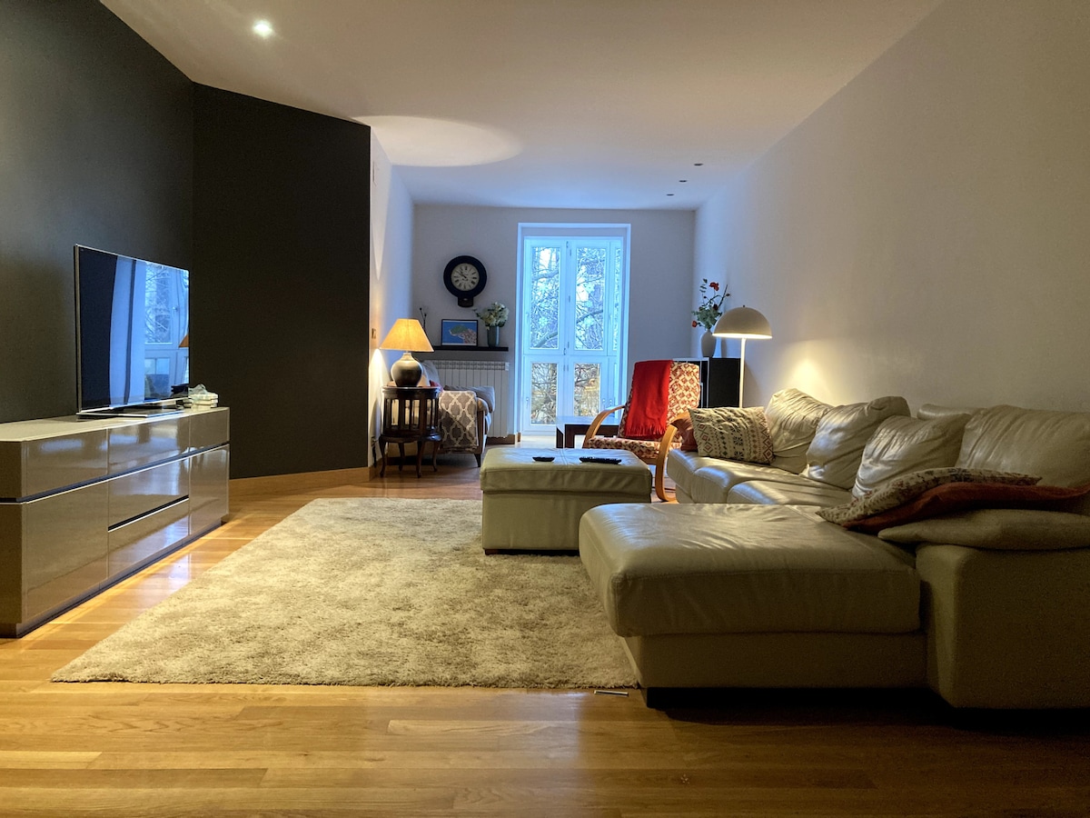 A spacious living area features a large sectional sofa in neutral tones, complemented by a contemporary wooden coffee table. A flat-screen television is positioned on a sleek, low-profile cabinet. Natural light filters through a large window, creating a warm atmosphere. Decorative plants and a lamp provide additional ambiance.