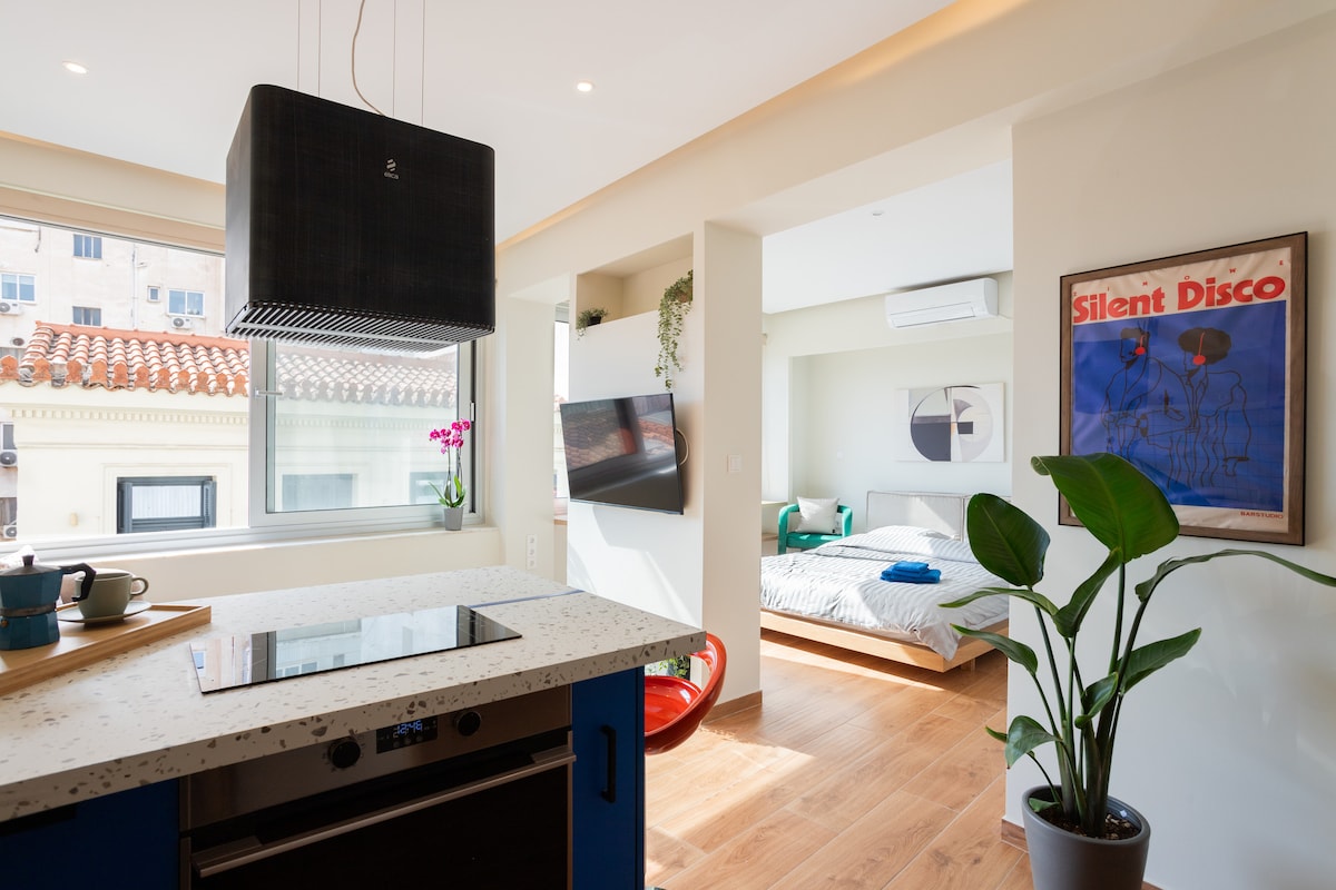 A modern kitchen area features a sleek countertop with a stovetop and small appliances. Natural light enters from a large window, highlighting a cozy living space and a plush bed in the background. Artwork adorns the wall, contributing a touch of personality to the design.