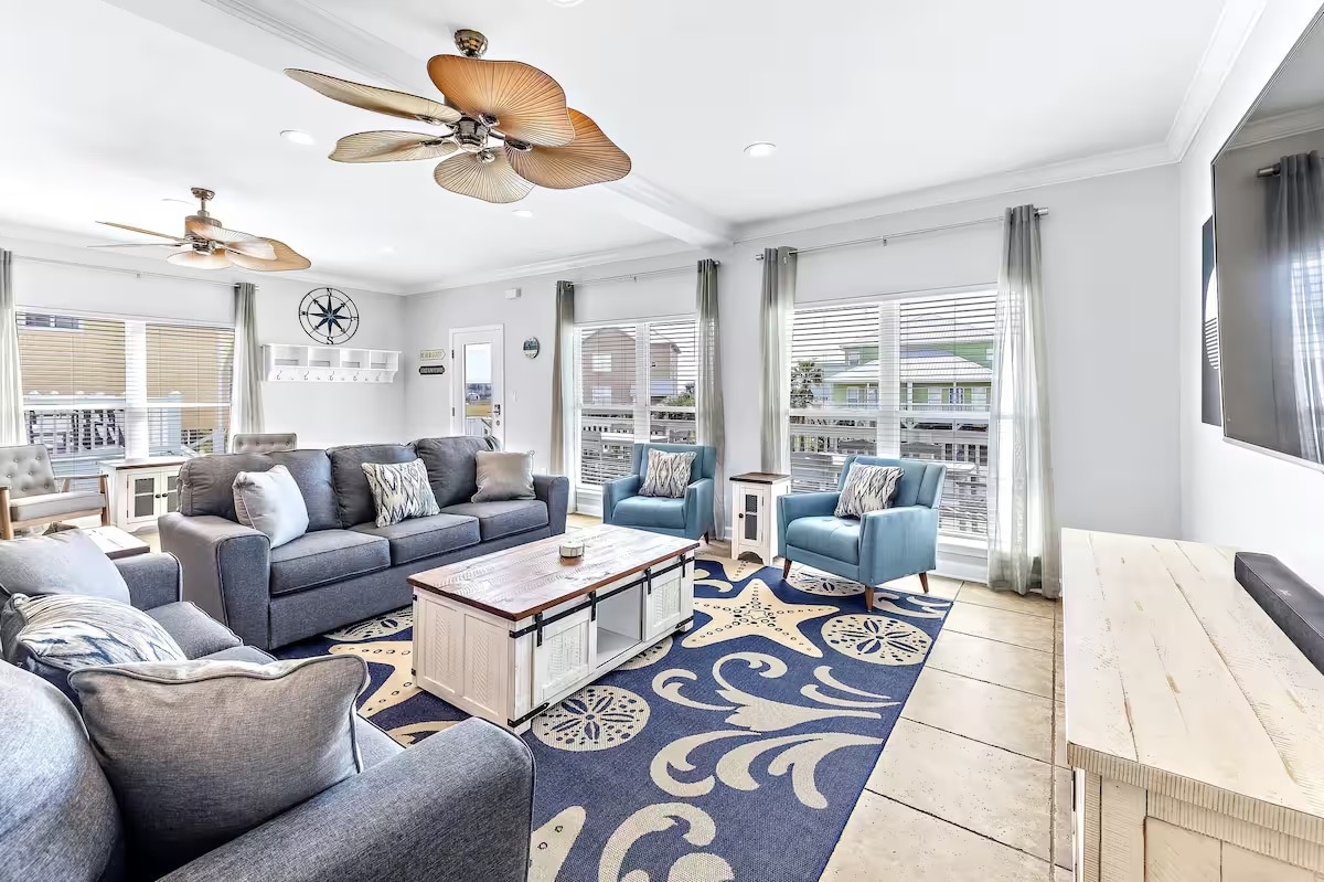 A spacious living area is furnished with a sectional sofa, accent chairs, and a coffee table. Large windows allow natural light to flood the room, revealing views of the outdoor area. A decorative area rug adds color and texture to the tiled floor.