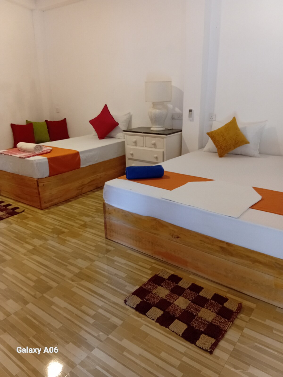 The sleeping area features two spacious beds, each adorned with colorful throw pillows. A bedside table holds a lamp, and the floor displays woven rugs. Natural light brightens the room, reflecting off the light-colored walls.