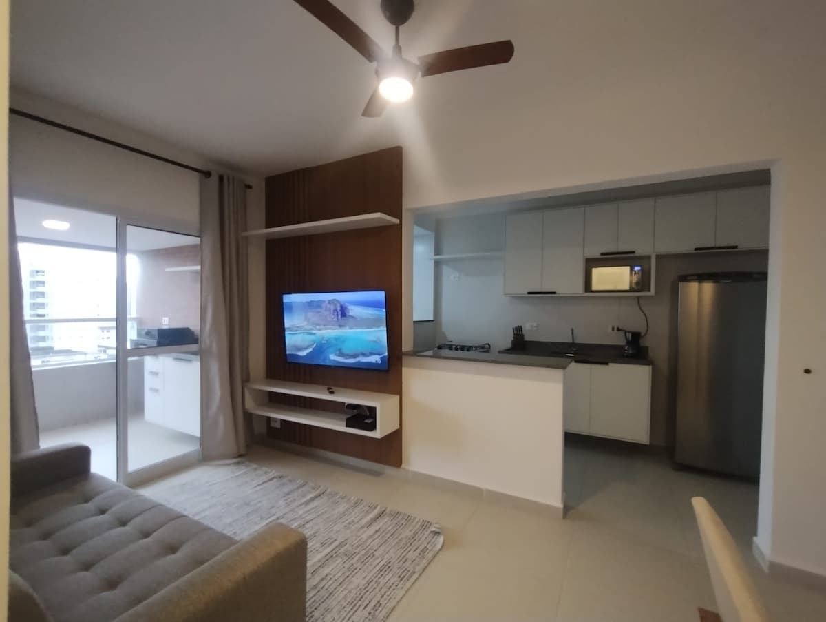 The living area features a comfortable sofa positioned near a mounted smart TV against a wooden accent wall. A ceiling fan is overhead, and natural light enters through a large window, leading to a balcony area.