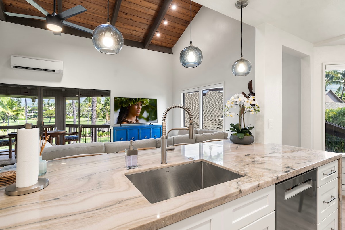 A bright kitchen space features a spacious countertop with a sink, complemented by stylish pendant lighting above. The room opens up to a living area with a large TV, showcasing views of the surrounding greenery through sliding glass doors.