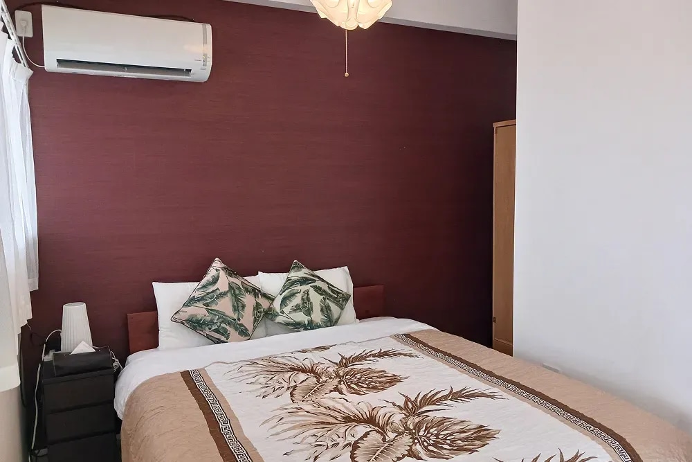 A spacious sleeping area features a large bed adorned with a decorative blanket and accented by tropical-themed pillows. Light filters through a nearby window, complemented by a softly lit bedside lamp and an air conditioning unit mounted on the wall.