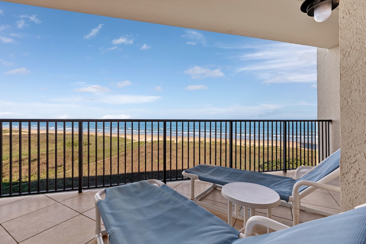 A spacious balcony features two chaise lounges beside a small round table, offering a view of the shoreline. The railing provides safety, while the sky above displays soft clouds against a bright blue background.