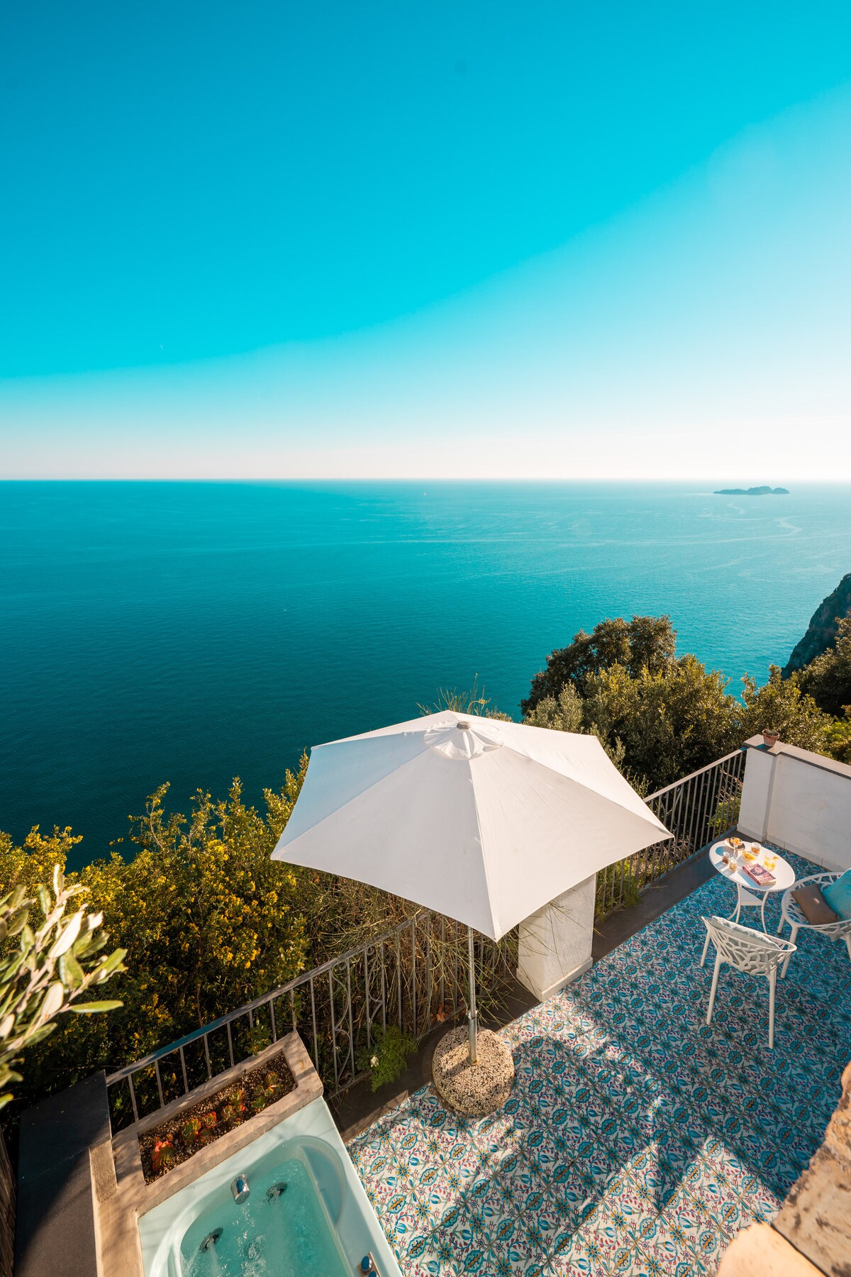 A serene terrace overlooking the sea features a jacuzzi surrounded by lush greenery. An umbrella shades a small table set for two, complemented by intricate tile work underfoot. The expansive view captures the tranquil blue ocean meeting a clear sky.