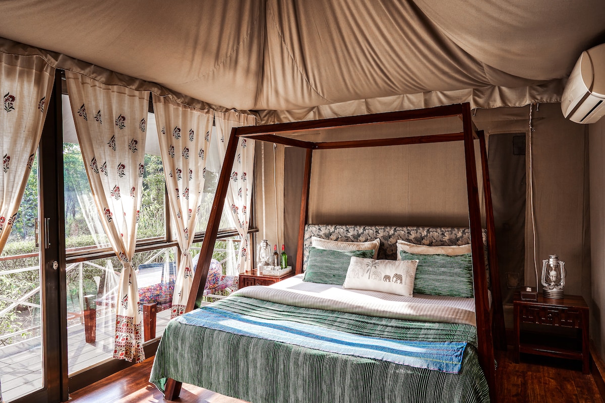 A spacious tented accommodation features a comfortable bed adorned with a decorative blanket. Large windows with patterned curtains allow natural light to filter in, complementing the wooden furnishings that enhance the rustic charm of the interior.