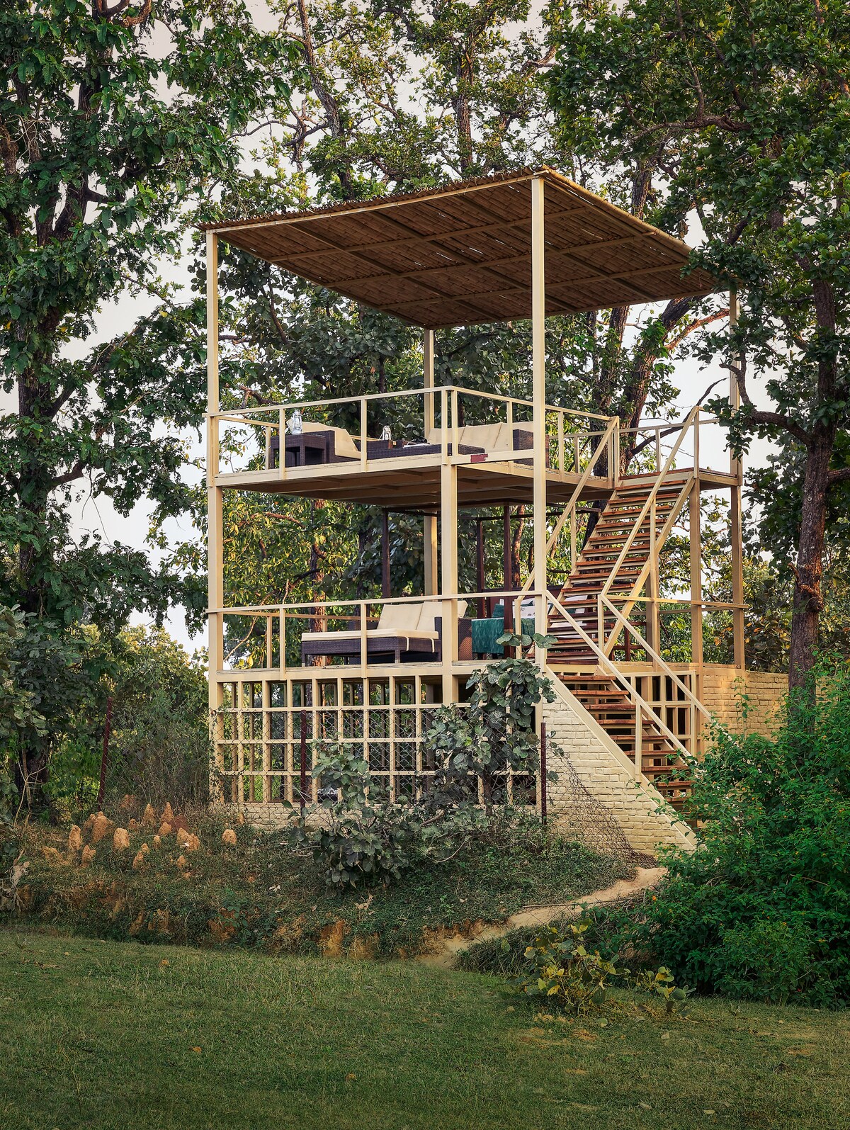 A two-story structure is viewed, elevated on stilts with an open deck on the upper level. Sturdy stairs lead up to the deck, surrounded by greenery. The building's design features open railings and is complemented by natural foliage, integrating it into the forest environment.