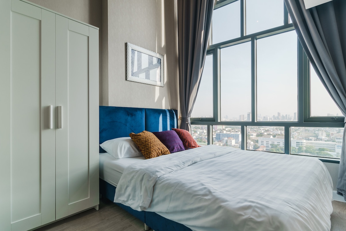 A bright bedroom is showcased, featuring a large window with city views. A spacious bed is adorned with decorative pillows and a light duvet. A wardrobe is positioned nearby, complementing the room's modern design. Soft light fills the space from the window.