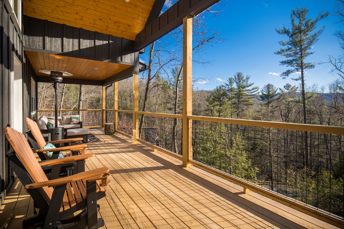 A spacious covered porch features wooden lounge chairs and a cozy sofa, offering a view of the surrounding trees and mountains. Natural light floods the area through large glass doors, creating a serene outdoor space for relaxation.