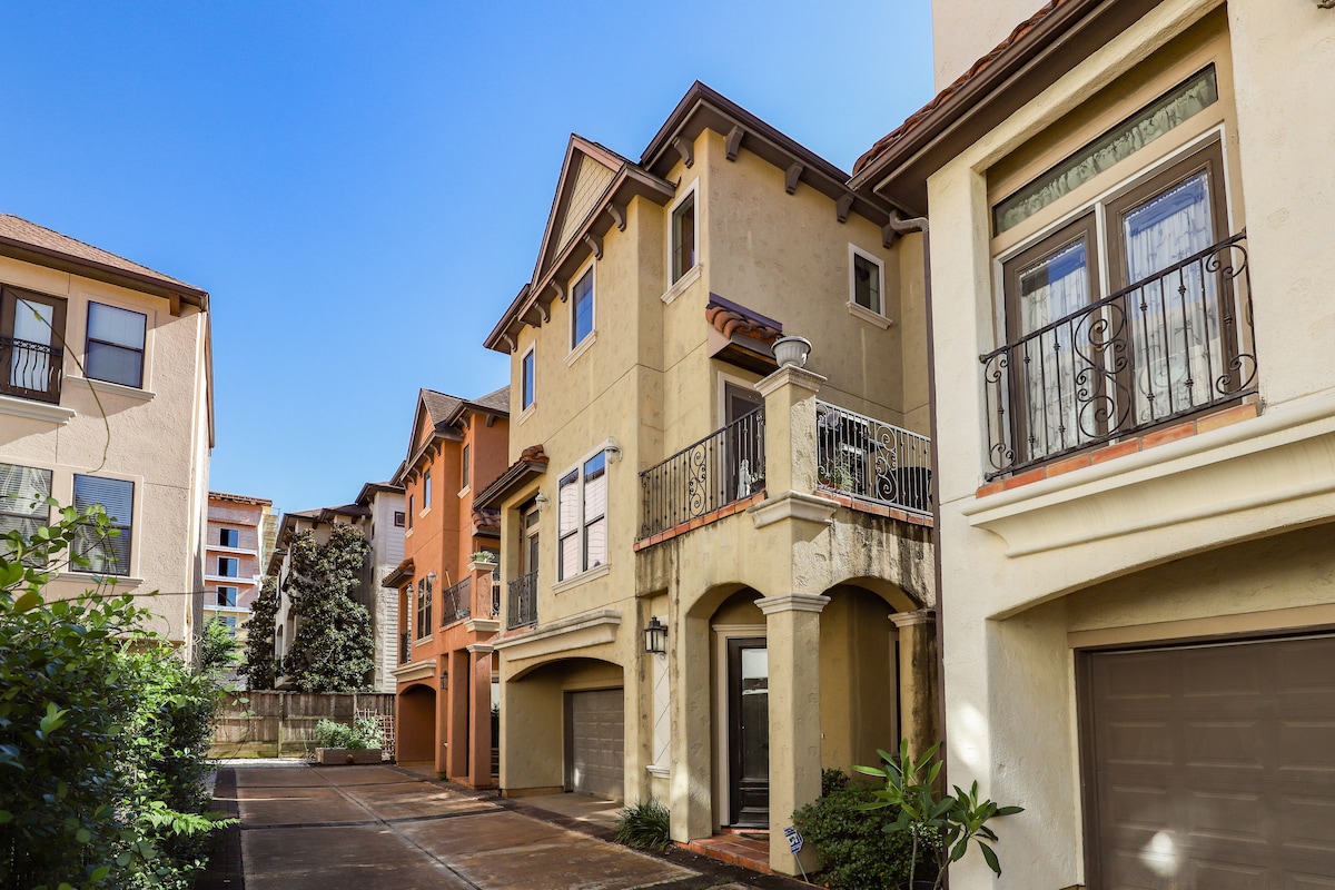 A row of multi-story homes with varied architectural styles is shown along a smooth driveway. Balconies are visible on the second floors, framed by decorative iron railings. Lush greenery is present at the edge of the driveway, complementing the vibrant facade of the homes.
