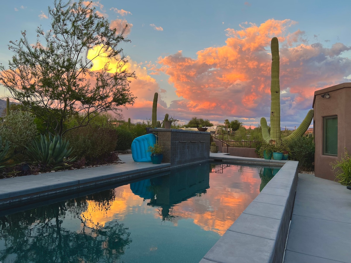 A 40-foot lap pool reflects the vibrant colors of the sunset, framed by a lush desert garden. Tall saguaros and other native plants create a serene backdrop against the sky, which features soft clouds transitioning from orange to pink.