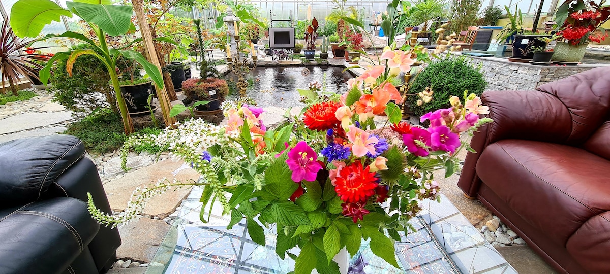 An indoor space is filled with lush greenery, featuring a pond surrounded by decorative plants. In the foreground, a vibrant floral arrangement encompasses a variety of colors and species. Comfortable seating is visible on either side, inviting relaxation amidst the natural decor.