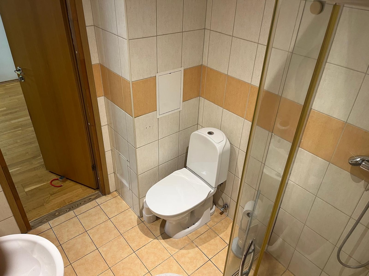 The bathroom features neutral tiles with a combination of light and orange accents. A modern toilet is positioned next to a glass shower enclosure. The space includes an ample area for movement, enhancing accessibility.