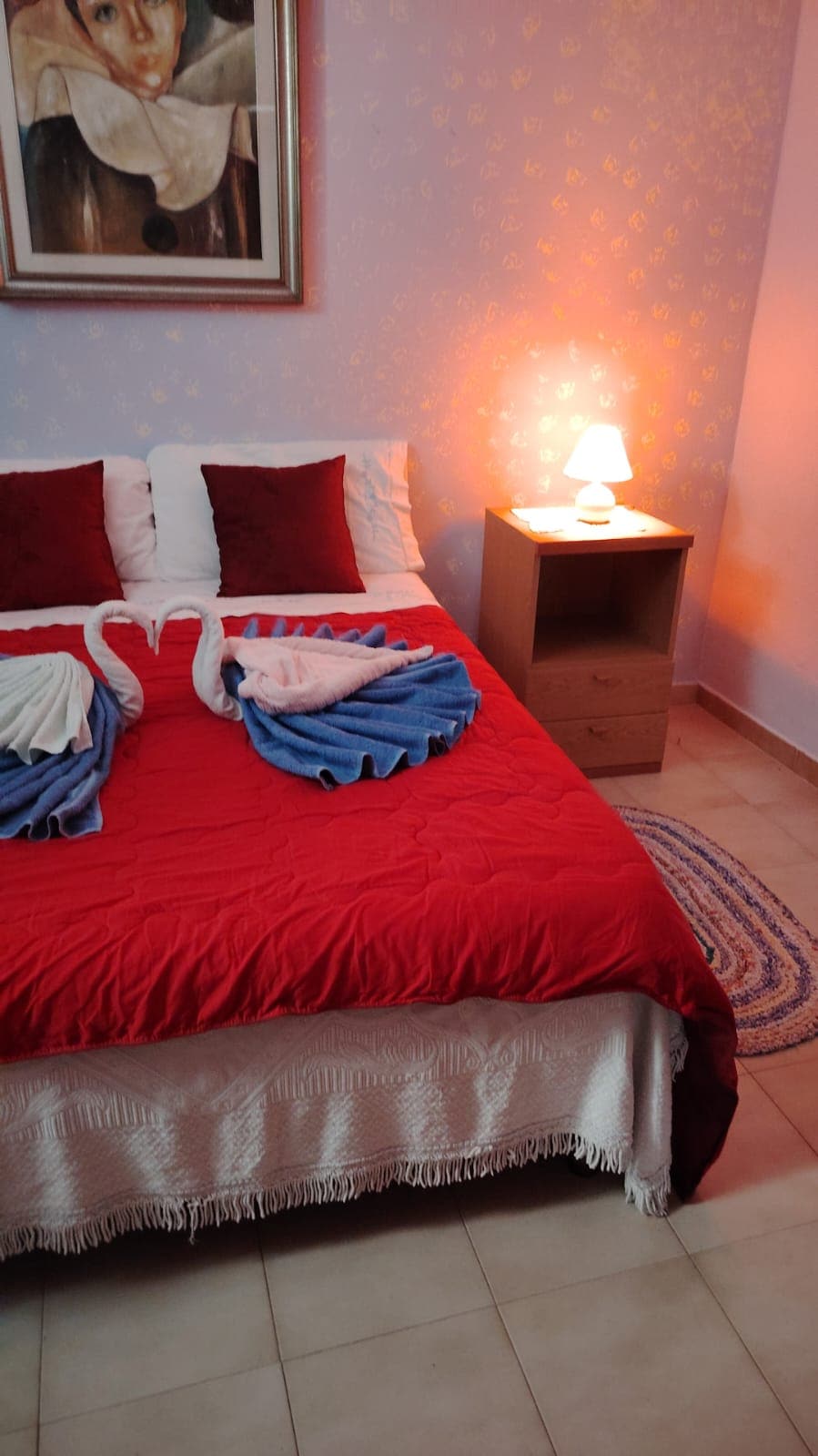 A spacious bed is adorned with a red blanket, complemented by decorative towels arranged in the shape of swans. A soft glow is provided by a bedside lamp, and a piece of wall art hangs above the bed, enhancing the room's warmth.