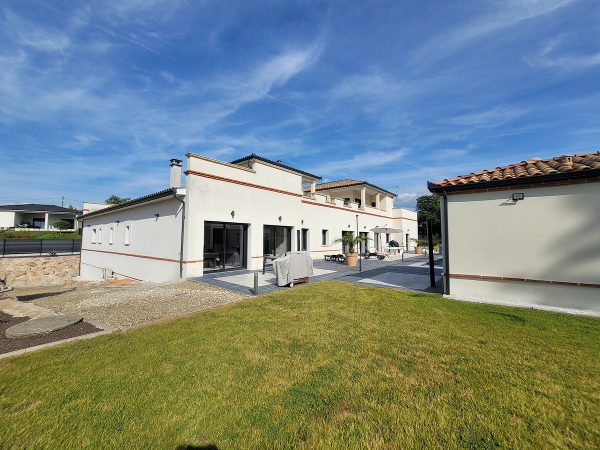 A modern, two-story building is viewed from the outside, showcasing expansive glass doors and a neatly landscaped lawn. The property is set against a clear blue sky, with outdoor seating visible on the patio, providing ample space for relaxation.