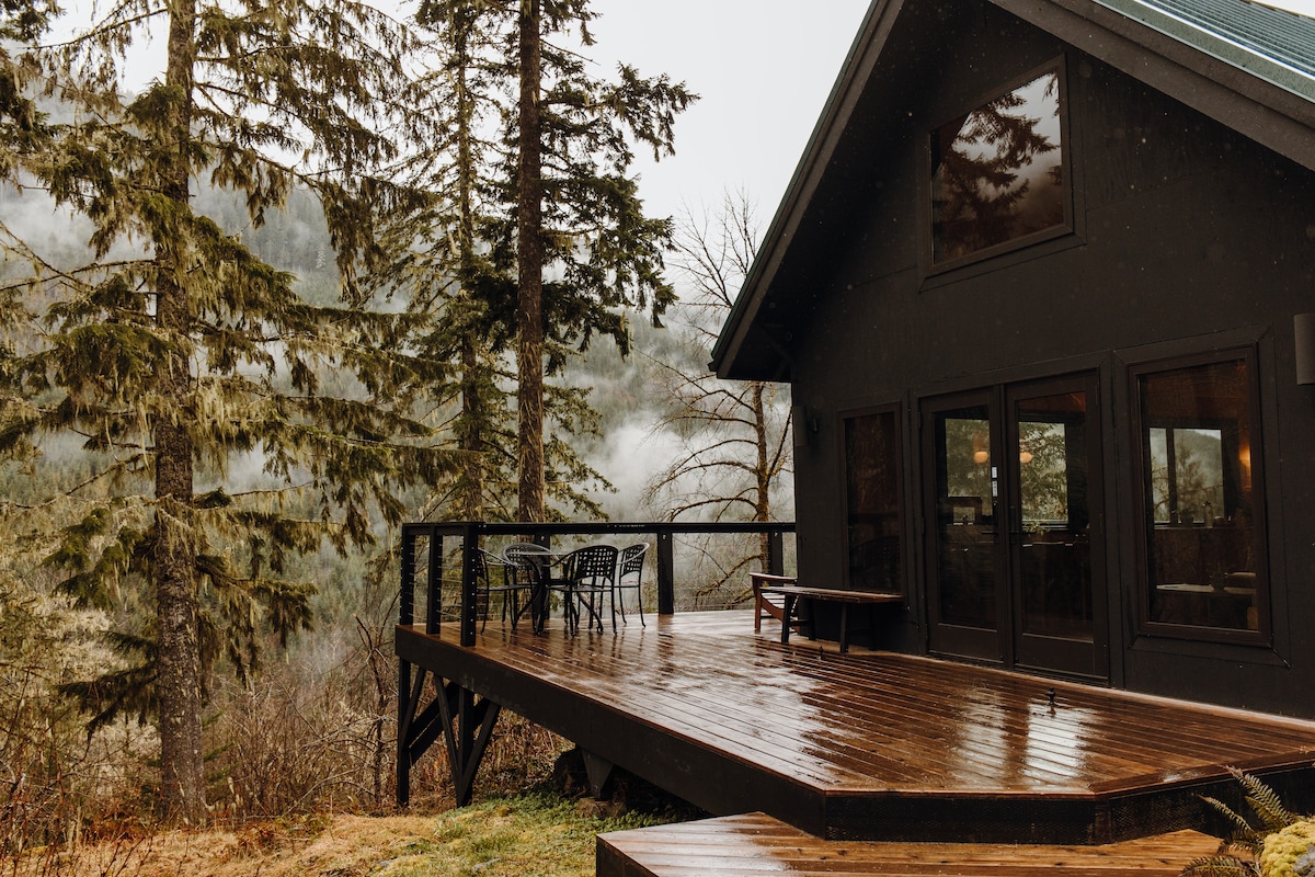 The home’s expansive outdoor deck features a dark wood surface, surrounded by tall evergreen trees. A wrought iron table with four chairs provides a spot for outdoor relaxation, while large windows allow for views of the misty valley beyond.
