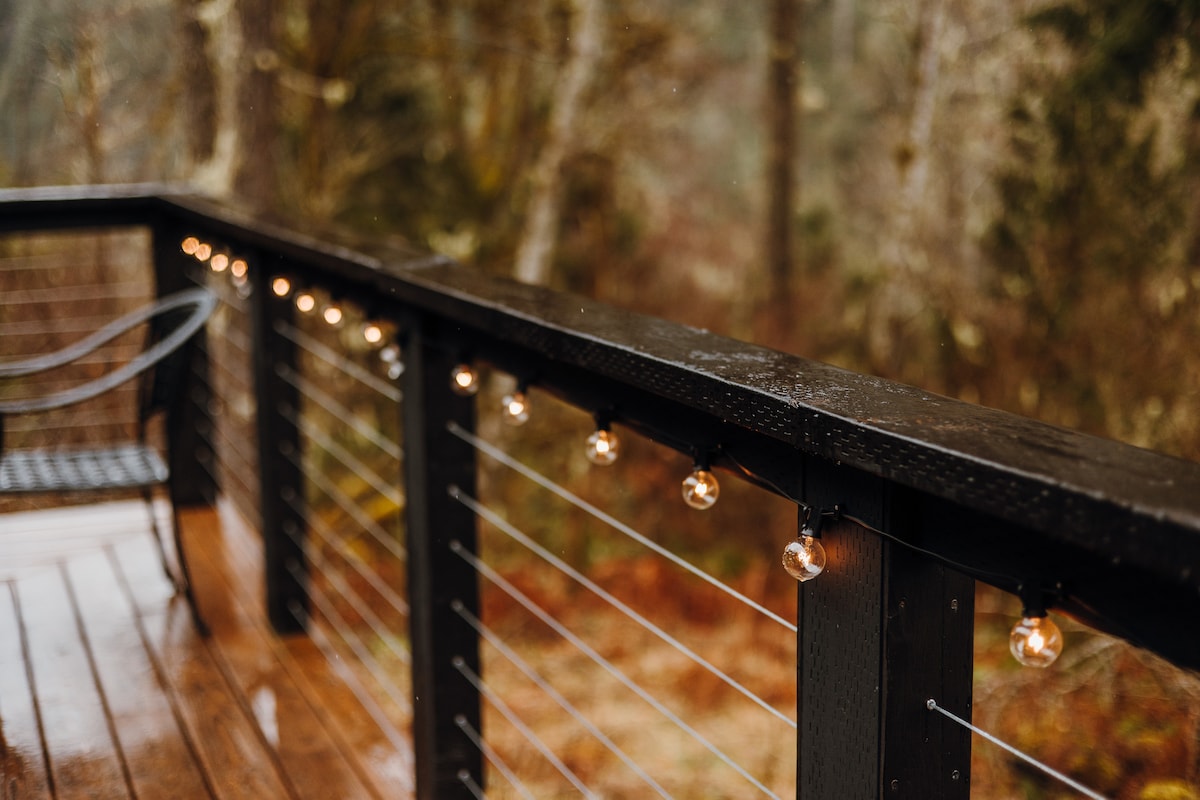 An outdoor deck railing is adorned with string lights, casting a soft glow. The surface is wooden, and a black chair is positioned on the deck. Dense trees and a misty landscape are visible in the background, enhancing the serene atmosphere.