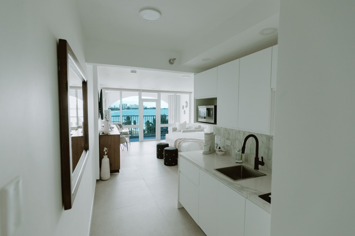 A modern studio space is shown, featuring a fully equipped kitchen with sleek white cabinetry. Light-colored tile flooring extends throughout. Expansive glass doors lead to the private balcony with ocean views. Natural light fills the room, creating an airy ambiance.
