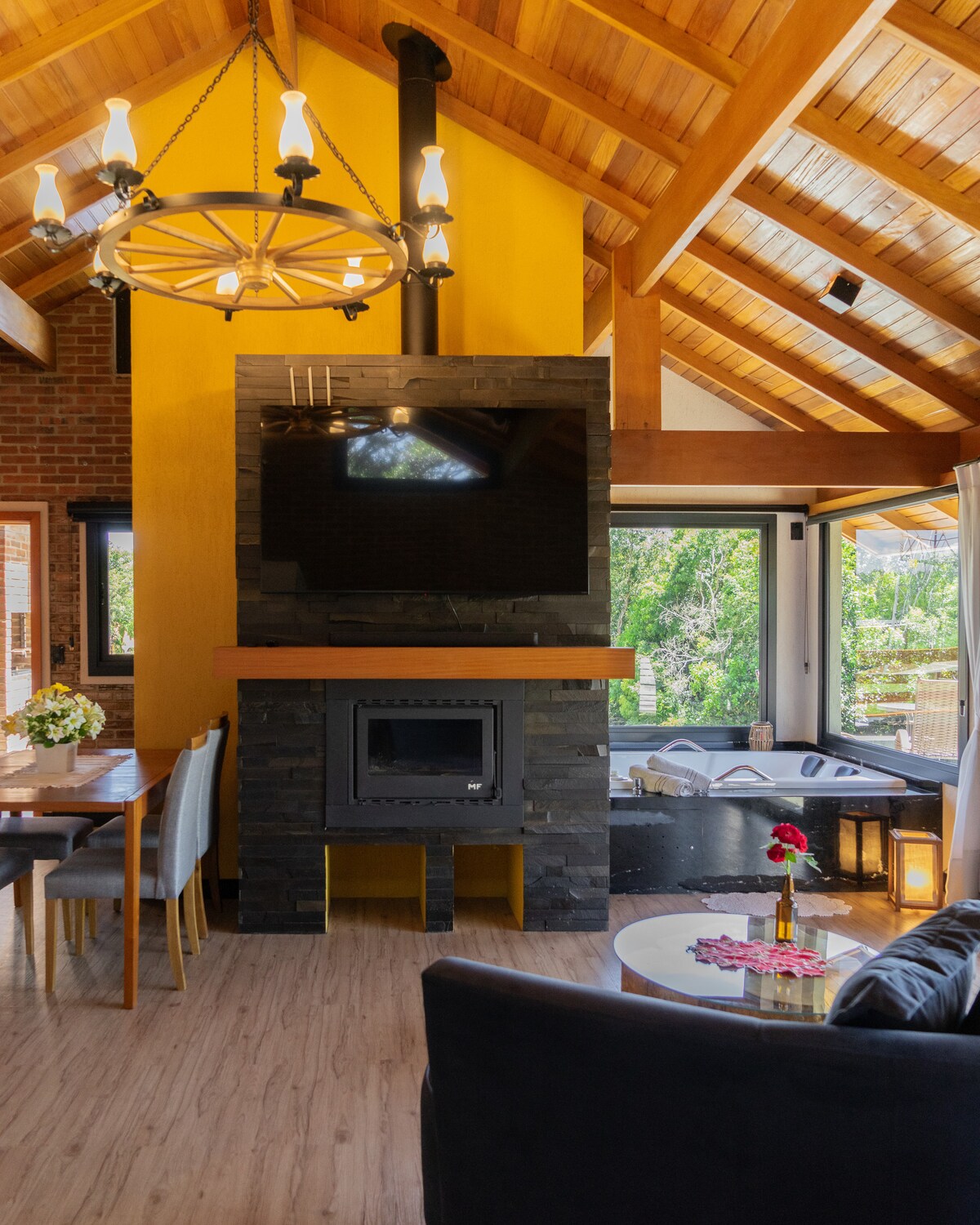 An open living space features high wooden ceilings and a stone fireplace, complemented by a large television mounted above it. A cozy seating area includes a dark sofa and a coffee table, while a dining table with chairs is positioned nearby. Light streams through large windows, revealing lush greenery outside.